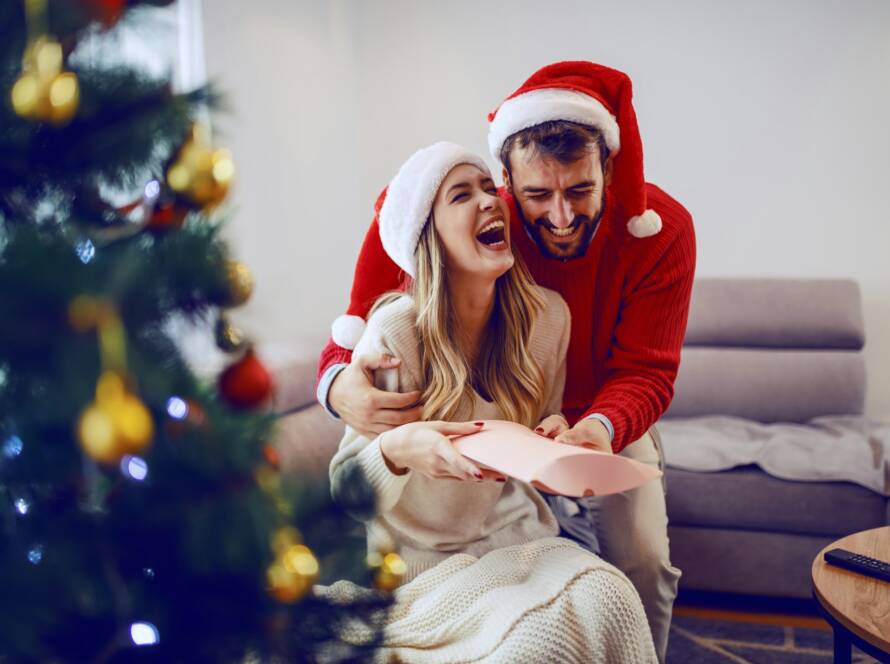 A couple wearing Santa hats laughing together while opening a gift, capturing the warmth and joy of the cozy season of love.