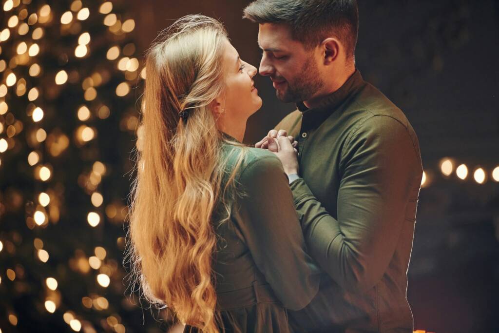 A romantic couple holding hands and looking into each other’s eyes with holiday lights glowing in the background, symbolizing the cozy season of love.