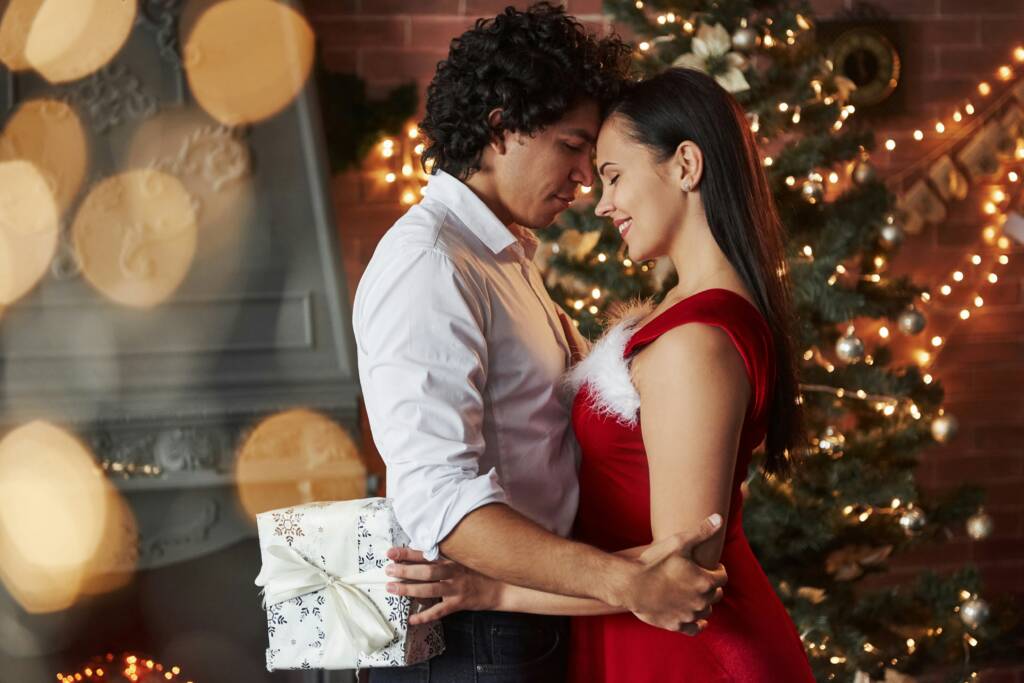 A couple embracing near a decorated Christmas tree while holding a wrapped present, reflecting the warmth of the cozy season of love.